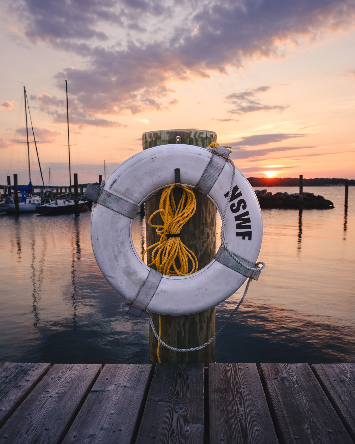 A life preserver attached to a wooden post on a dock at sunrise. The background features moored sailboats and calm water reflecting the colorful sky.