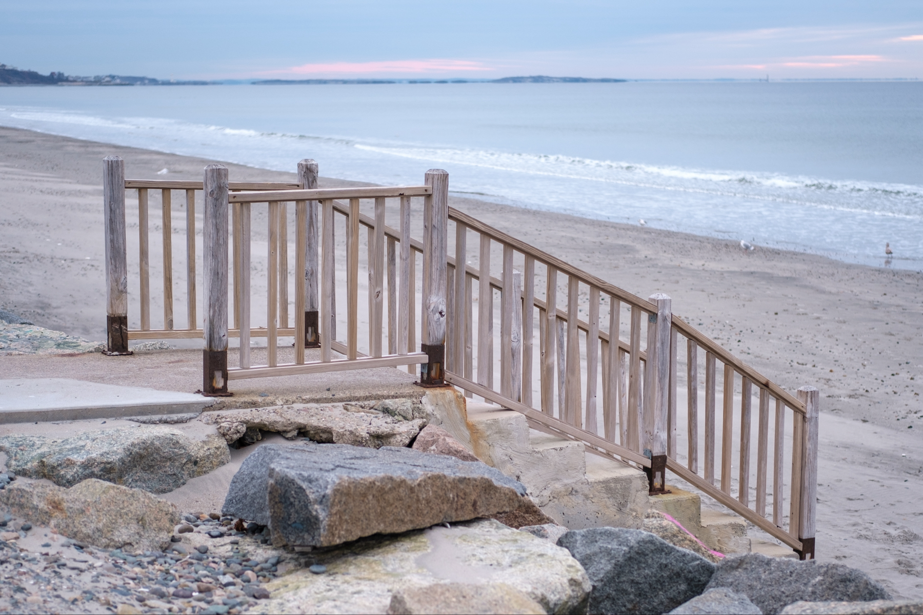 A wooden stair railing leading down to a sandy beach with rocks in the foreground and the ocean in the background a pale sunrise. 