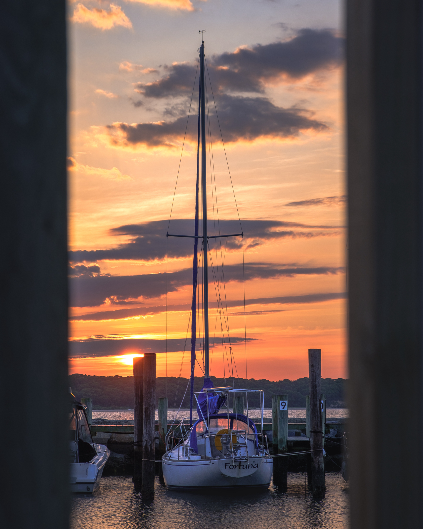 A sailboat docked at a marina during a sunrise, framed by two vertical wooden posts. The boat’s name “Fortuna” is visible on the stern. The sky is painted with warm hues of orange and pink, with purple clouds scattered above