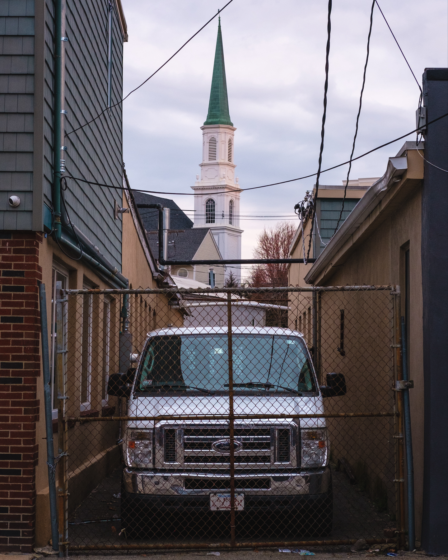A white van parked in an alley with a chain-link gate, framed by buildings on either side, and a church spire visible in the background.