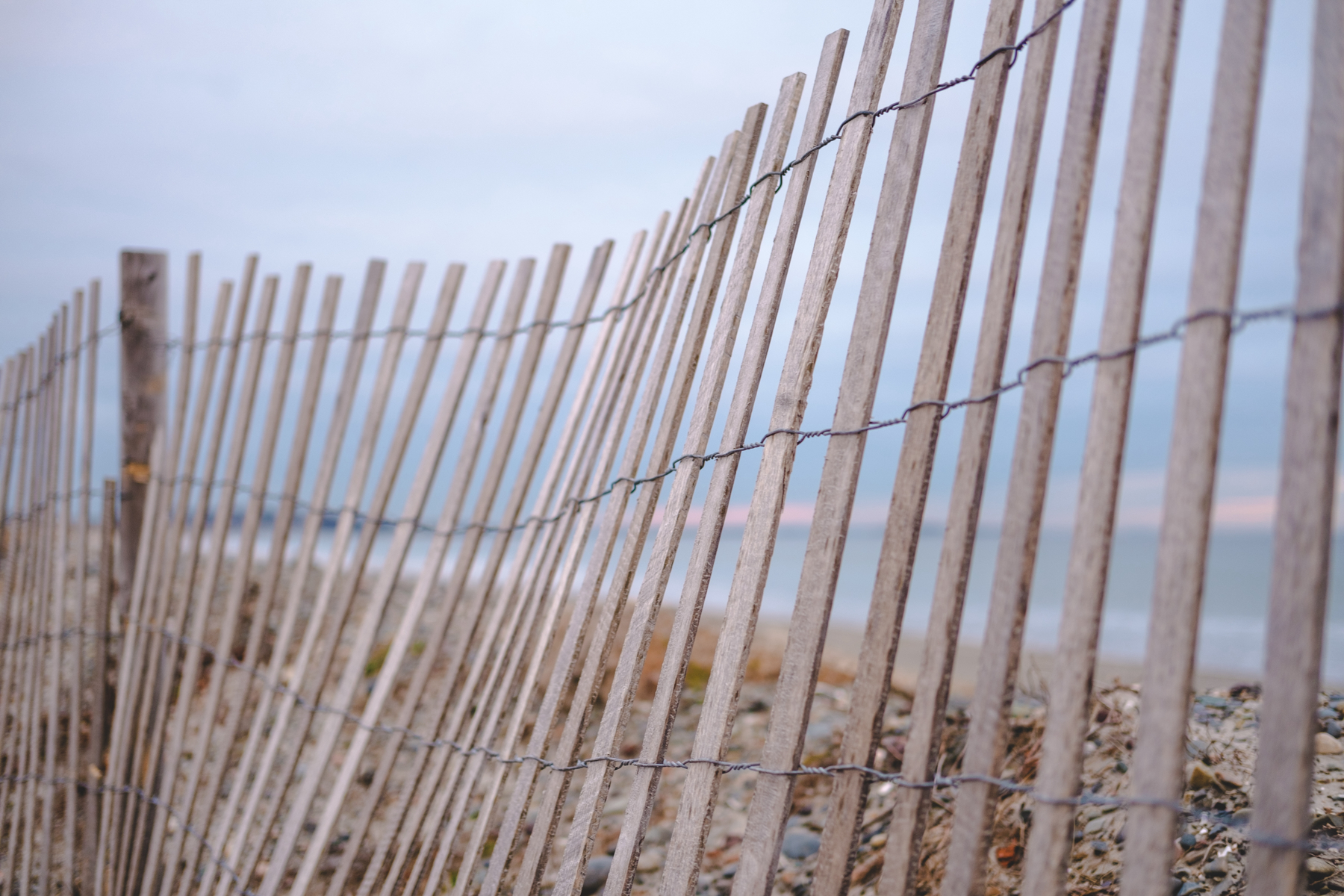 A wooden slat fence along a beach with blurry ocean and pale sunrise sky in the background.